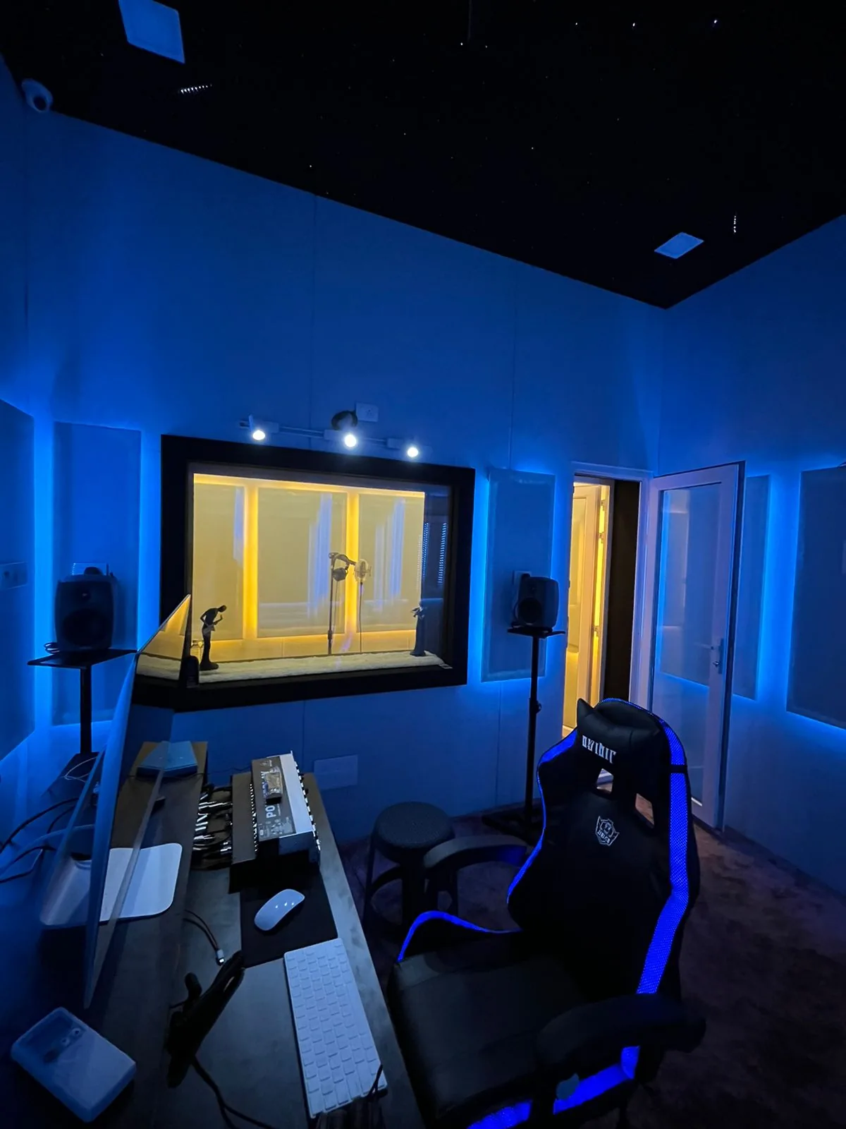 A recording studio control room in blue light, looking through the observation window into the vocal booth